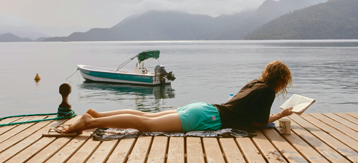  A serene outdoor scene featuring a person relaxing by water, reading a book, with a peaceful natural backdrop.